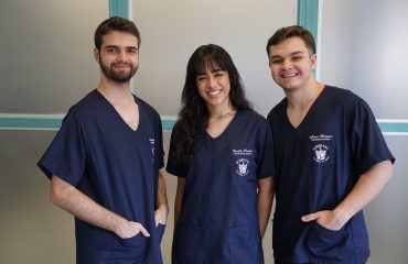 Dentistas recém-formados como alunos da soberana. nas extremidades dois rapazes e no meio uma menina. Todos trajam pijama cirúrgico na cor azul marinho