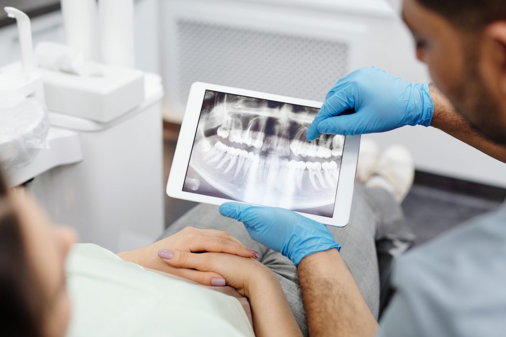 A imagem foca no uso da tecnologia no atendimento odontológico. O paciente está deitado na cadeira de atendimento com as mãos cruzadas sobre o ventre, enquanto as mãos do dentistas estão vestidas com luvas azuis, mostrando um exame de imagem em uma tela de tablet