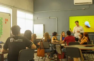 A imagem mostra uma sala de aula na melhor faculdade de Odontologia de Juiz de Fora, a Soberana. Em primeiro plano, os alunos virados de costas prestando atenção no professor, que aparece no fundo direito da imagem.
