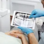 A imagem foca no uso da tecnologia no atendimento odontológico. O paciente está deitado na cadeira de atendimento com as mãos cruzadas sobre o ventre, enquanto as mãos do dentistas estão vestidas com luvas azuis, mostrando um exame de imagem em uma tela de tablet