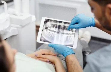 A imagem foca no uso da tecnologia no atendimento odontológico. O paciente está deitado na cadeira de atendimento com as mãos cruzadas sobre o ventre, enquanto as mãos do dentistas estão vestidas com luvas azuis, mostrando um exame de imagem em uma tela de tablet